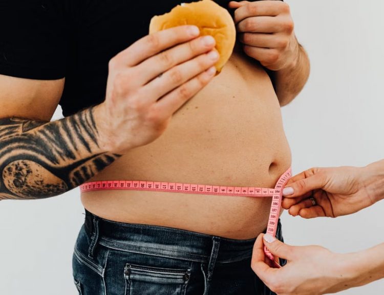 A man with tattoos holding a burger as another person measures his waistline with a tape measure.