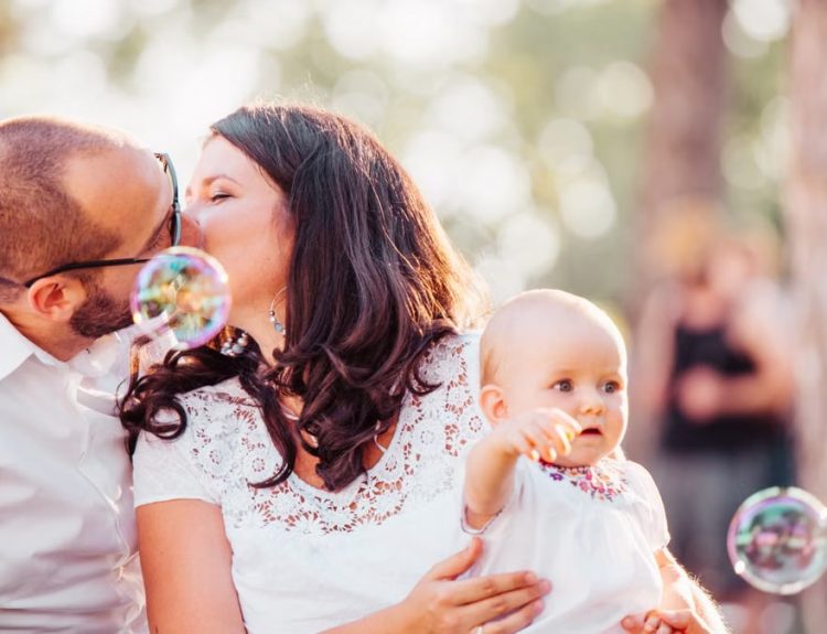 A tender family moment captured with a couple kissing and a baby holding bubbles outdoors.